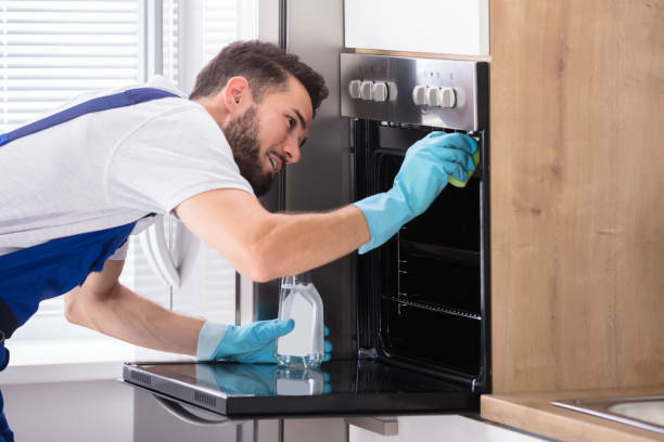 Happy Male Janitor Cleaning Oven With Sponge In Kitchen